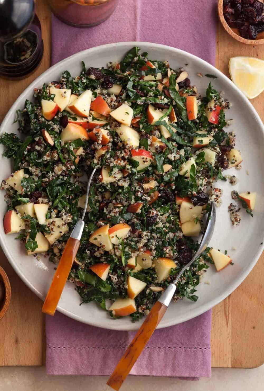 Kale and quinoa salad made with crisp apple, dried cranberries, almonds and Pecorino on plate with serving spoons