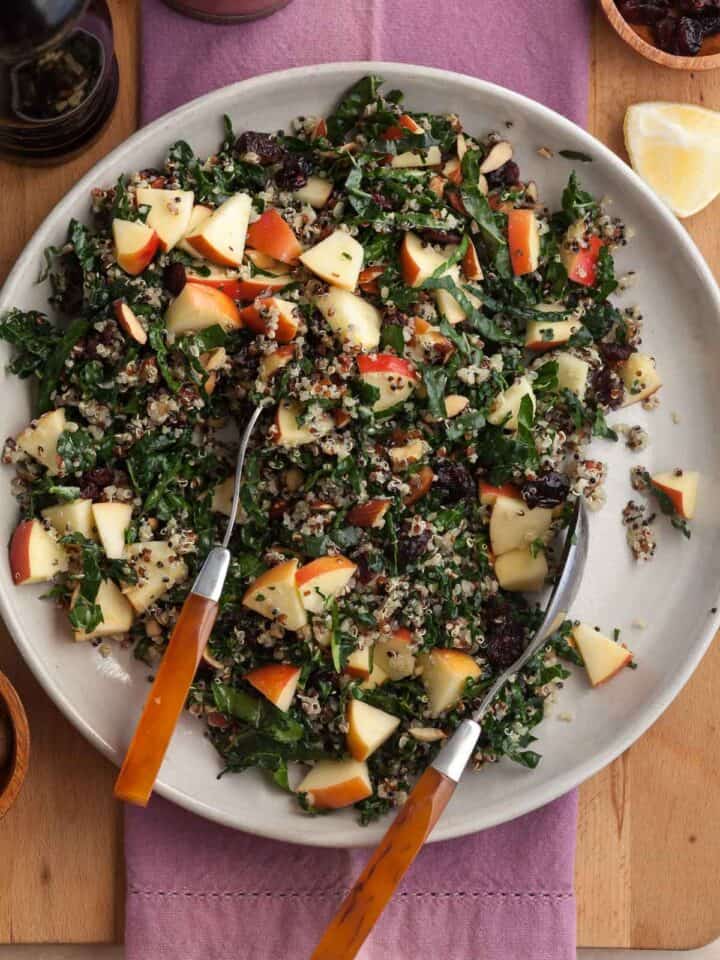 Kale and quinoa salad made with crisp apple, dried cranberries, almonds and Pecorino on plate with serving spoons